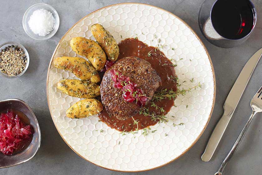 Beef tenderloin, onion jam, red wine mustard sauce, and thyme on a white plate with fingerling potatoes, and a glass of red wine.