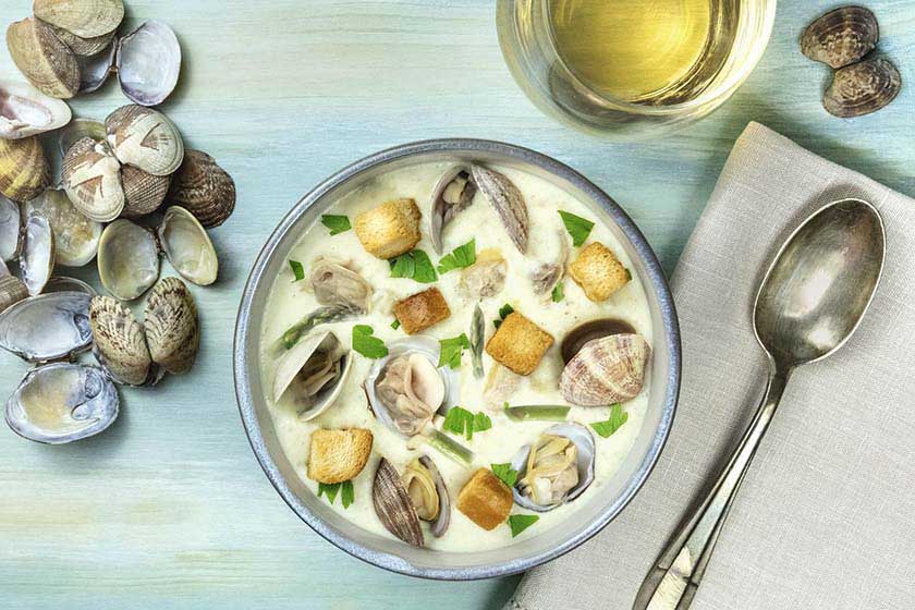 Bowl of creamy asparagus chowder with clams in their shells, sprinkled with parsley and paired with a glass of white wine.