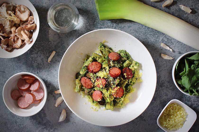 Bowl of mushroom and sausage pasta with kale pesto surrounded by recipe ingredients