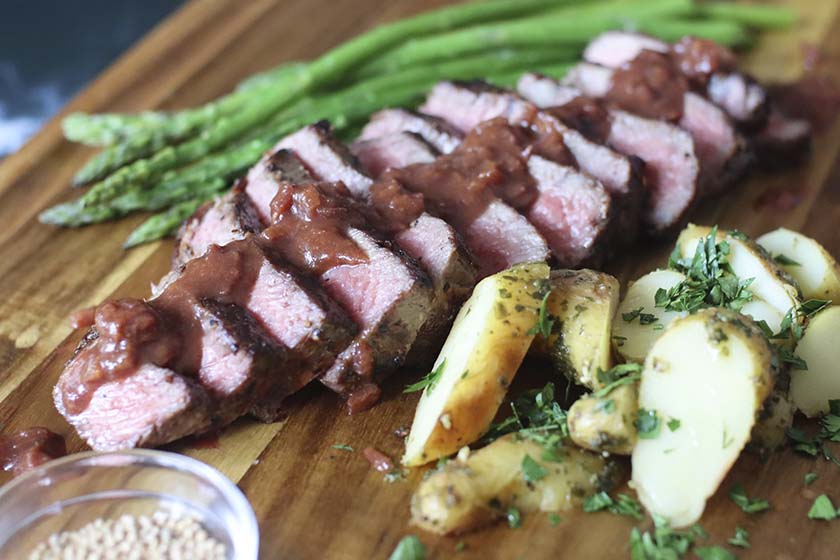 Sliced NY steak drizzled with Sauce Robert on a wood plank with roasted potatoes and asparagus