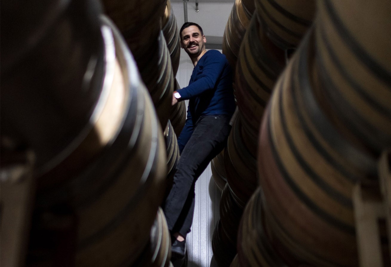 A smiling Sam Baron, winemaker for Kivelstadt Cellars, standing high between a narrow row of stacked wine barrels