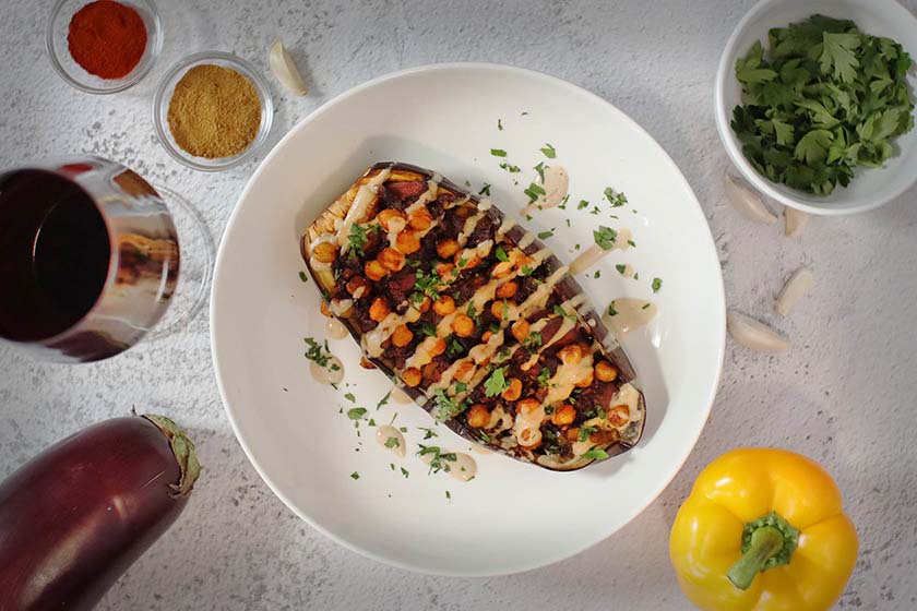 Stuffed eggplant drizzled with a maple, balsamic, and tahini sauce. Its surrounded by recipe ingredients and paired with a red wine.