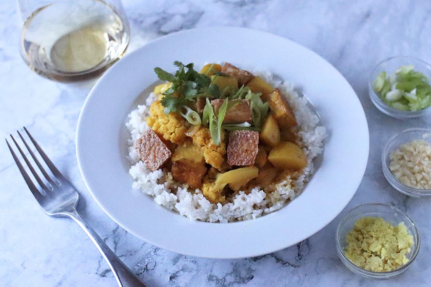 Bowl of potato and cauliflower veggie coconut curry surrounded by ingredient dishes and a glass of white wine