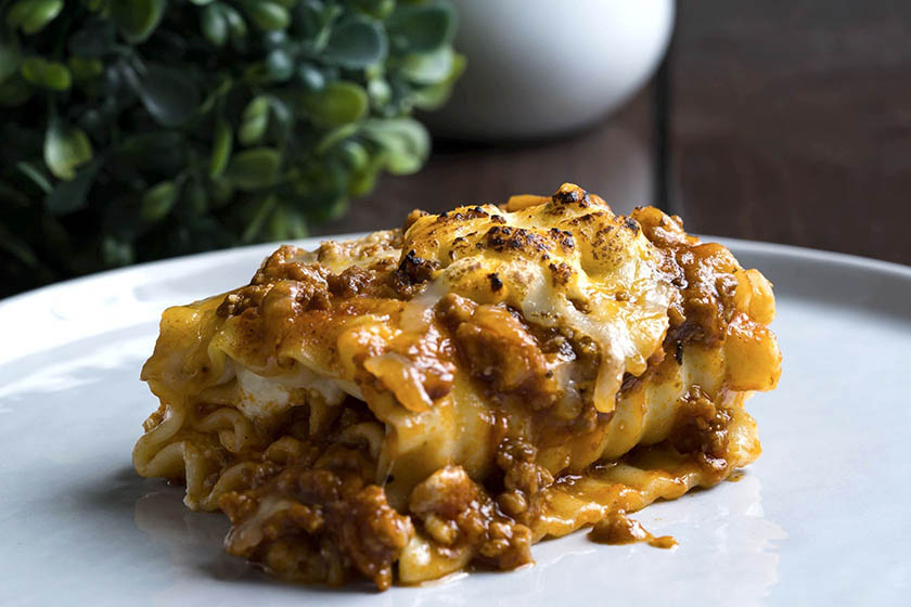 A slice of lasagna bolognese on a white plate topped with cheese